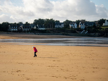 A Child In A Red Jacket Walks On The Sandy Bay Beach At Low Tide. In The Background Houses On The Rocky Shore Are Visible.
