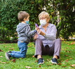 © progat - Grandmother and little kid playing with a smartphone in the backyard while wearing protective masks due to coronavirus outbreak