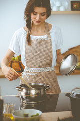 Wall Mural - Young brunette woman cooking soup in kitchen. Housewife holding wooden spoon in her hand. Food and health concept