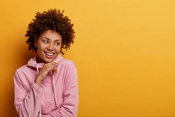 Wall Mural - People, lifestyle concept. Positive dark skinned female happy to find good opportunities for future work, holds chin looks away with broad smile hears wonderful news feels upbeat, poses on yellow wall
