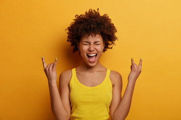 Positive joyful woman makes heavy metal sign, has fun on music rocking festival, exclaims loudly, closes eyes, gestures actively, dressed in casual wear, isolated over yellow wall. Rock n roll baby