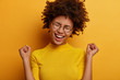Cheerful triumphing woman achieves victory, raises clenched fists with triumph, rejoices winning prize, dressed casually, keeps eyes closed, isolated over yellow background. Celebration concept