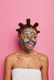 Fototapeta  - Vertical shot of cheerful African American woman has fresh clean skin, looks aside, applies clay mask, enjoys facial treatment, stands wrapped in bath towel, isolated on pink wall. Beauty face care