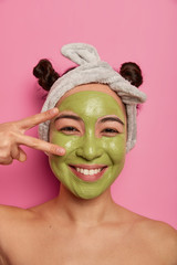 Wall Mural - Close up shot of natural Asian woman has fun during beauty procedures, makes peace sign over eye, applies green purifying facial mask, cleanses skin, stands with naked body isolated on pink background