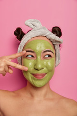 Wall Mural - Pretty dreamy young female model makes peace gesture over eye, applies nourishing green mask on face, wears headband, stands naked indoor, enjoys beauty treatments, isolated on pink studio wall