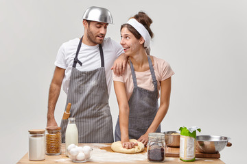 Wall Mural - Positive housewife gives culinary master class for husband, shows how to make and knead dough, prepare breakfast together in cozy home, make cookies, wear aprons, spend free time at kitchen.