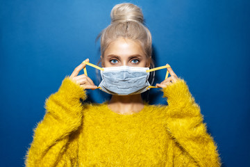 Wall Mural - Studio portrait of young girl with blonde hair bun, wearing medical face mask against coronavirus and yellow sweater, on textured background of phantom blue color.