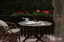 Wooden Brown Table On The Terrace Of The House In The Garden. On The Table Are A Laptop, A Cup Of Coffee, Flowers, A Candle, A Clock, Lipstick, Perfume. In The Background Are Green Trees And Petunia 