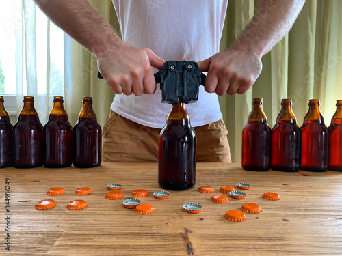 Cuadros en Lienzo Craft beer brewing at home, man closes brown glass beer bottles with plastic capper on wooden table with orange crown caps