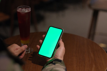 Wall Mural - Over shoulder of man holding green screen smart phone in the bar at night. A glass of cocktail on woode table. Blur background