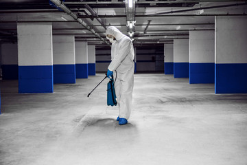 Full length of worker in protective sterile suit and mask disinfecting underground garage from corona virus / covid-19.