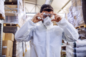 Young man in protective suit putting protective glasses and preparing himself for disinfection from corona virus / covid-19. Warehouse interior. Warehouse is full of food products.