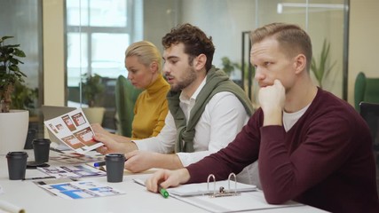 Wall Mural - Tracking right shot of male chief designer presenting printouts with website interface templates for different devices to colleagues sitting at table in team meeting