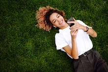Young Woman Sitting On The Grass  In A City Park And Using A Smartphone. Technology And Modern Lifestyle Concept.