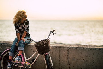 Wall Mural - People enjoying outdoor leisure actvity looking a beautiful coloured sunset - beautiful adult young woman with long curly blonde hair  enjoy the relax after a bike tour - horizon and sun with ocean