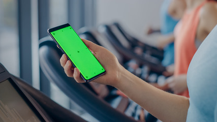 Athletic Woman Does Gestures while Using Green Screen Smartphone and Running on the Treadmill in a Gym. Fit Female Posts on Social Media, Takes Pictures, Watches Videos, Exercises in Fitness Club