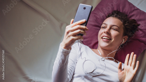 Young happy woman using mobile phone for video call , gesturing hi to friends, relatives or parents. Beautiful girl making facetime video calling with smartphone lying on the bed.