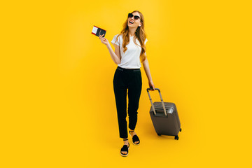 Happy beautiful woman, with a suitcase and passport with tickets on a yellow background. The concept of travel, business