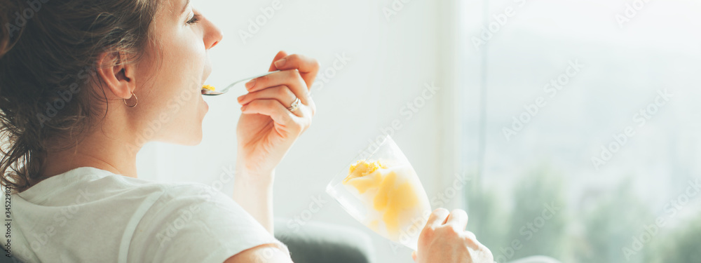 Young woman in white t-shirt sitting near big bright window eating ice cream. Wide screen, panoramic - obrazy, fototapety, plakaty 