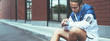 © Alex from the Rock - Young athlete in blue windrunner sitting on the street adjusting running program for his morning workout using smart watches on his hand. Wide screen, panoramic