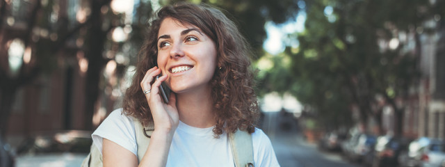 Curly woman talking on the mobile phone. Walking along the street with backpack and traveling. Wide screen, panoramic