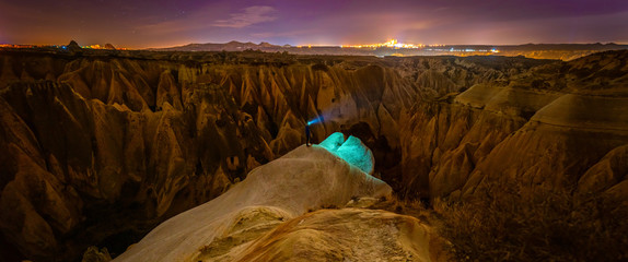 Wall Mural - Panoramic view of red valley in Cappadocia at night with stars in the background and person in the middle surounded by rock formations. Astrophotography in Turkey. Background blank space image.