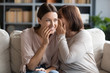 Middle aged mother whispering in smiling adult daughter ear, gossiping, sharing secret, happy beautiful woman hearing good news from mature mum, sitting together on couch, trusted relations