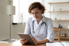 Serious Professional Doctor Wearing White Coat And Stethoscope Holding Modern Touchscreen Gadget Using Digital Tablet Computer At Work. Healthcare Tech Medical Data Network And Telehealth Concept.