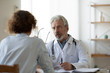 © fizkes - Professional senior male physician consulting female patient in hospital. Old doctor examining young adult woman at medical visit. Senior man therapist talking to client at healthcare checkup meeting