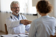 © fizkes - Friendly smiling professional physician handshaking female patient at medical consultation. Happy old male doctor greeting woman client shake hands express trust and gratitude for healthcare service.