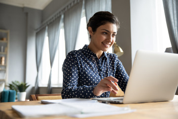 Head shot smiling pretty indian girl sitting at table, looking at laptop screen. Happy hindu ethnic woman reading message email with good news, chatting with clients online, working remotely.