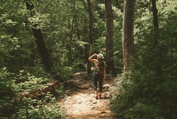 woman walks in the summer forest with a backpack and a hat. stands and looks at the opening of sunli