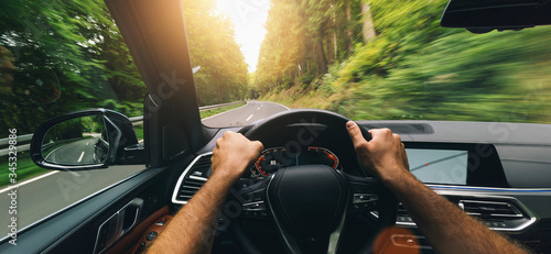 Obraz Dibond Ręce kierowcy na kierownicy, szybki samochód w wiosenny dzień na wiejskiej drodze, dobra zabawa podczas jazdy pustą autostradą w trakcie wycieczki - widok z perspektywy pierwszej osoby POV