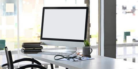 Computer monitor with white blank screen putting on doctor working desk together with stack of books, stethoscope, digital thermometer, clipboard, potted plant, glasses, mouse and keyboard
