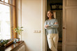 © shurkin_son - Beautiful casually dressed female pensioner posing indoors standing by windowsill with house plants, looking through window with pensive facial expression, leaning against white wall, daydreaming