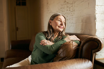 Wall Mural - Indoor image of beautiful pensive sixty year old female with gray hair sitting comfortably in stylish leather sofa by window, having thoughtful dreamy facial expression, wants to have walk outside