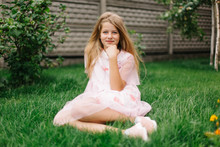 Beautiful Blue-eyed Girl With Long Blond Hair. Little Girl In A Pink Flamingo Dress. Summer Bright, Emotional Photo.  Girl Model With Long Hair Sits On Green Grass