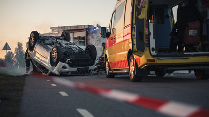 On the Car Crash Traffic Accident Scene Rollover Smoking Vehicle Lying on its Roof in the Middle of the Road after Collision. In the Background Medics Perform First Aid, Fire Engine