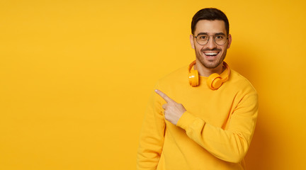 Horizontal banner of trendy man looking amazed and surprised pointing to copy space on left with finger, isolated on yellow background