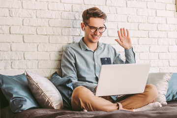 Stylish young hipster man student in glasses waving hand to laptop while talking online video call and sitting on couch. Happy businessman freelancer having web meeting, conference. Work at home