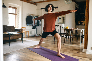 Full length image of handsome young male with strong athletic body practicing yoga indoors, standing in warrior 2 pose or virabhadrasana, taking deep breaths. Sports, active lifestyle and vitality