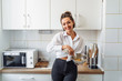 © Daria - Beautiful, attractive, smiling young woman kneading dough with whisk for banana pancakes in steel dishes in a kitchen with white, light interior. Monochrome photo without bright accents