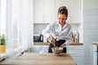 © Daria - Beautiful, attractive, smiling young woman kneading dough with whisk for banana pancakes in steel dishes in a kitchen with white, light interior. Monochrome photo without bright accents