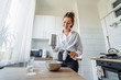 © Daria - DIY home cooking concept. A bright brunette woman sifting the flour for pancakes through a sieve, salting the dough, adding milk. Young woman smiling, loves to cook in a beautiful modern kitchen.