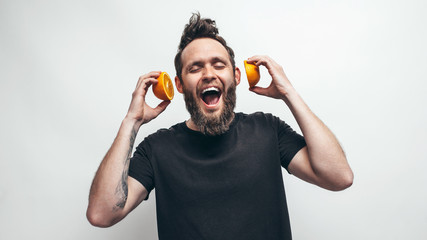 Young bearded man holding orange slices in hands in front of his ears like headphones for listening to music. Healthy eating