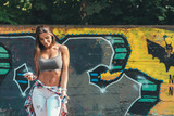 Fototapeta Młodzieżowe - Relaxed young confident stylish woman tying shirt around waist on graffiti wall background