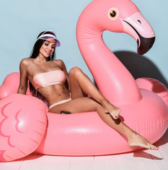 Poster - Young tanned slim brunette woman in sexy pink beach bikini and cap sitting on big pink flamingo and smiling