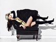 © Dmitry Lobanov - Young brunette woman in golden swimsuit, cap, black high boots and jacket lying on black velvet vintage armchair and looking down