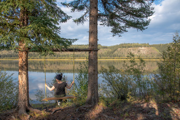 Woman swinging on rustic, hand made swing on the side of a river in summer. 