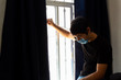 © Antonio Tanaka - young man wearing a blue medical mask looking out of a window very sad about the coronavirus quarantine